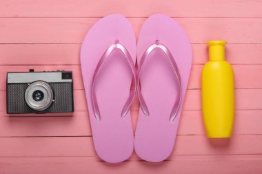 Plaj tatili konsepti, yaz geçmişi. Güneş kremi şişeli parmak arası terlikler, pembe ahşap arka planda kamera. Üst görünüm