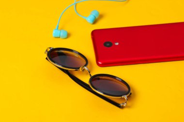 Sarı arka planda kadın aksesuarları ve aletleri. Kulaklıklar, zarif yuvarlak güneş gözlükleri, sarı arka planda akıllı telefon.
