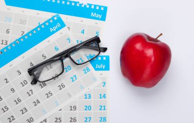 Aylık takvim kağıtları, gözlükler, beyaz arka planda kırmızı elma. Ekonomik hesaplama, maliyet. Üst görünüm