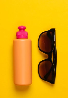 Sarı arka planda güneş gözlüklü bir şişe güneş kremi. Plaj Tatili Konsepti. Üst görünüm