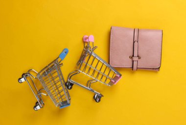 Sarı arka planda cüzdanı olan iki mini market arabası. Alışveriş konsepti. Üst Manzara. Minimalizm