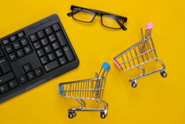 İnternetten alışveriş. Süpermarket arabalı bilgisayar klavyesi, sarı arka planda gözlükler. Üst Manzara. Düz yatıyordu 