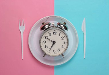 Piknik zamanı. Tek kullanımlık çatallı bir tabakta çalar saat ve pembe mavi pastel arka planda bıçak. Üst görünüm