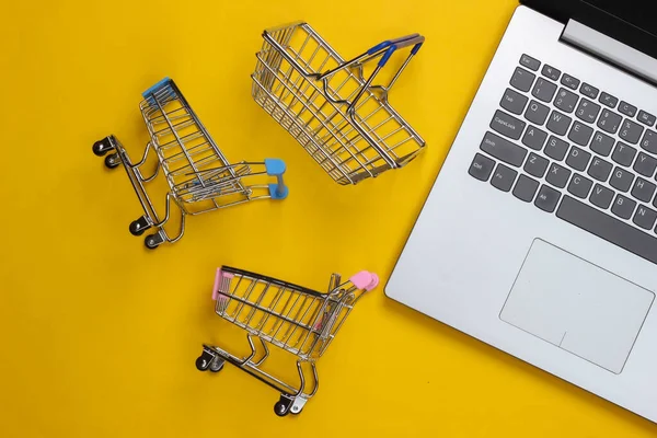 Online shopping. Laptop with supermarket trolleys, basket on a yellow background. Top view. Flat lay