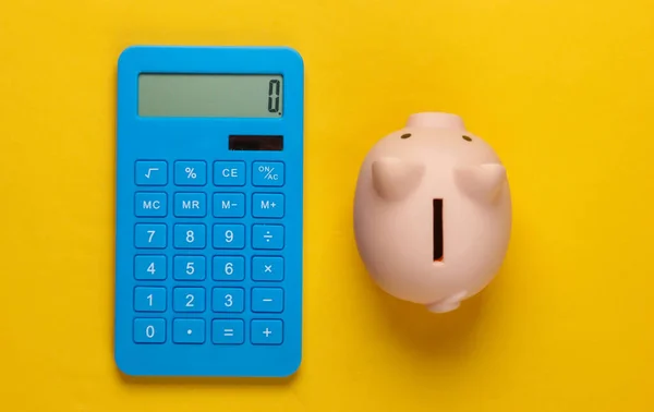 Piggy bank and blue calculator on yellow  background. Minimalistic studio shot. Overhead view. Flat lay.