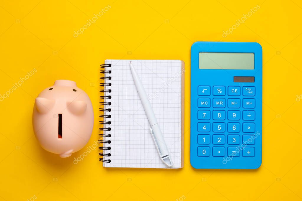 Piggy bank, notebook and calculator on yellow background. Minimalistic studio shot. Overhead view. Flat lay.