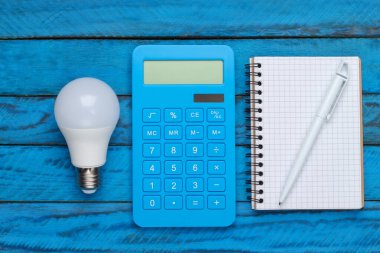 Elektrik tasarrufu. Önde gelen ampul ve hesap makinesi, mavi ahşap arka planda bir defter. Üst görünüm