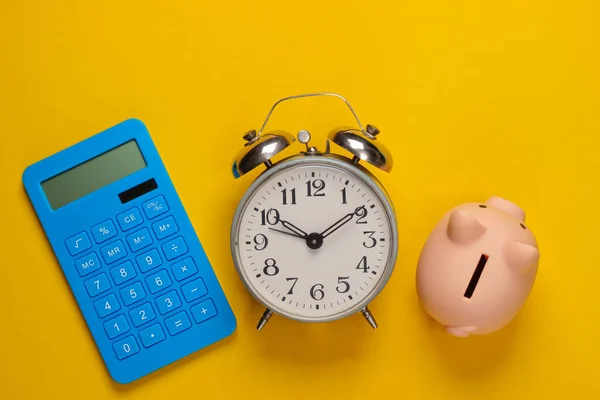 Piggy bank and alarm clock, blue calculator on yellow background. Minimalistic studio shot. Overhead view. Flat lay.