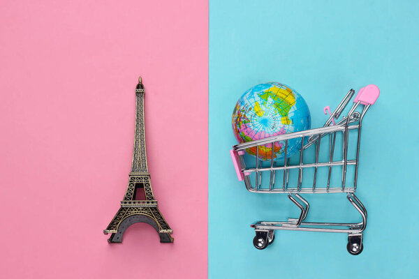 Минимализм путешествия и покупки натюрморт. Фигура Эйфелевой башни, глобус, тележка супермаркета на розово-голубом пастельном фоне. Вид сверху