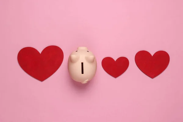 Piggy bank and hearts on pink pastel background. Minimalistic studio shot. Overhead view. Flat lay.