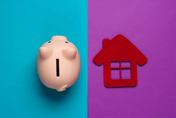 Save for housing. Piggy bank and house figure on purple blue background. Minimalistic studio shot. Overhead view. Flat lay.