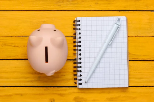 Piggy bank with notebook on a yellow wooden background. Top view