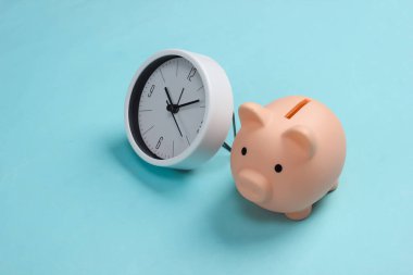 Yatırım yapma zamanı. Mavi pastel arka planda beyaz saat ve domuz kumbarası. Minimalist stüdyo çekimi.