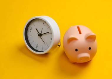 Yatırım yapma zamanı. Sarı arka planda beyaz saat ve kumbara. Minimalist stüdyo çekimi.