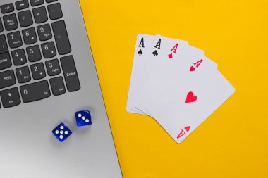 Sarı arka planda dört as, zar ve dizüstü bilgisayar klavyesi. Online poker kumarhanesi. Oyun bağımlılığı. Üst görünüm