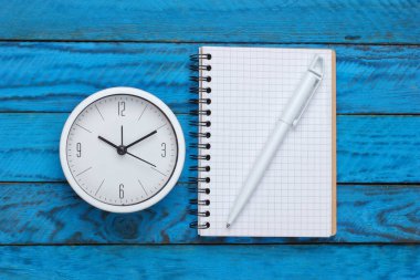 Mavi ahşap arka planda beyaz saat ve defter. Minimalist stüdyo çekimi. Üst görünüm