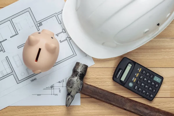 White construction safety helmet and piggy bank with blueprint, hammer, calculator on wooden boards. Building idea. House building costs. Top view