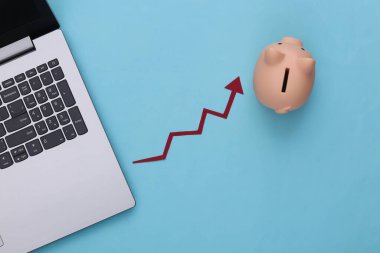 Laptop, domuz kumbarası, mavi arka planda kırmızı ok. Ok grafiği yükseliyor. Üst görünüm