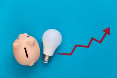 Enerji ve para tasarrufu kavramı. Büyüme oku mavi arka planda ampulle ve domuz kumbarasıyla birlikte yükseliyor. Üst görünüm