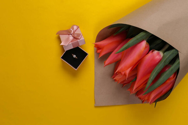 Marriage proposal. Bouquet of tulips, golden ring with diamond in gift box on yellow background. Top view