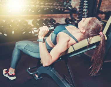 Spor kıyafetli genç bir kadın spor salonunda göğüs ve omuzların kasları için halterlerle egzersiz yapıyor.