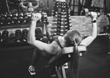 Spor salonundaki eğimli bankta göğüs ve omuzların kasları için halterlerle egzersiz yapan genç bir kadın. Arkadan bak. Siyah ve beyaz