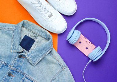 Retro tarzı. 80 'ler. Popüler kültür. Minimalizm. Denim ceket, kulaklıklar, ses kaseti, mor-turuncu arka planda beyaz spor ayakkabılar. Üst görünüm