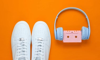 Retro tarzı. 80 'ler. Popüler kültür. Minimalizm. Ses kaseti olan kulaklıklar, turuncu arka planda beyaz spor ayakkabılar. Üst görünüm