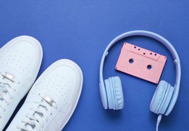 Retro tarzı. 80 'ler. Popüler kültür. Minimalizm. Kulaklıklar ve ses kaseti. Mavi arka planda beyaz spor ayakkabılar. Üst görünüm
