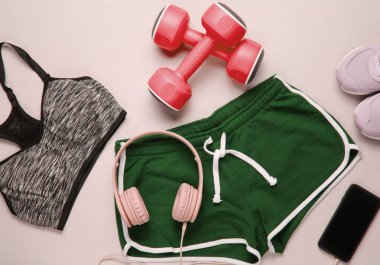 Gri arka planda spor kıyafetleri ve spor malzemeleri. Kulaklıklı akıllı telefon, plastik halterler, spor sutyen, şort, gri arka planda spor ayakkabılar. Üst görünüm
