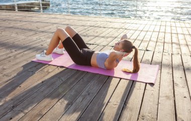 Spor kıyafetli genç bir kadın gün doğumunda plajda paspasın üzerinde yatarken karın kaslarını çalıştırıyor.