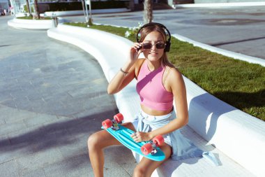 Şık giyinmiş, güneş gözlüklü, kaykaylı ve kulaklıklı genç sarışın kadın yaz güneşli bir günde açık havada.