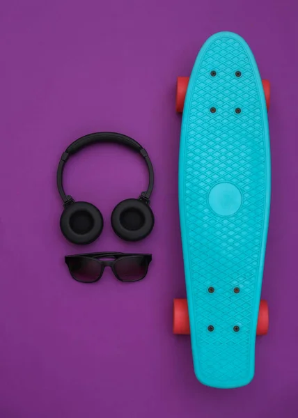 Genç hippi aksesuarlarının düz kompozisyonu. Kruvazör tahtası, kulaklık ve mor arka planda güneş gözlüğü. Üst görünüm