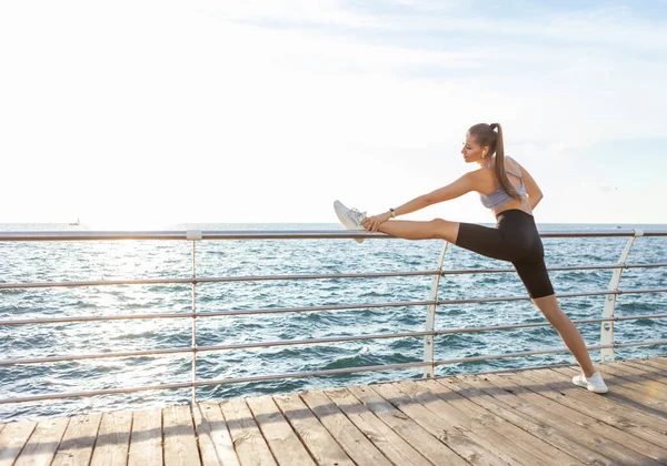 Mükemmel vücutlu zayıf formda bir kadın gün doğumunda sahilde antrenmandan önce bacak esneme egzersizi yapıyor.