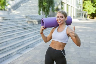 Neşeli formda kadın yoga minderini tutuyor ve dışarıda antrenman yapmaya hazırlanıyor. Sağlıklı yaşam tarzı kavramı