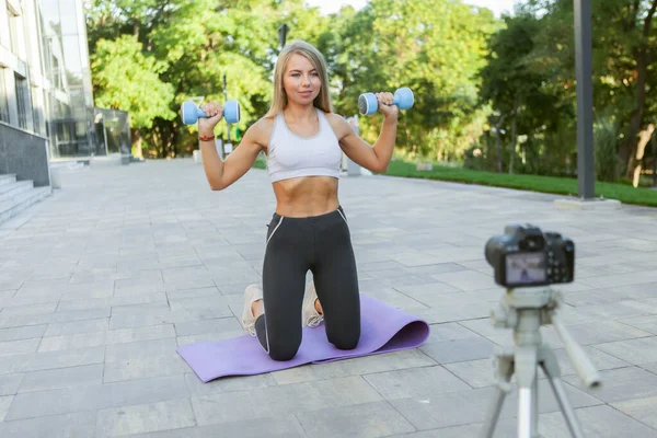 Fitness, sports and video blogging concept. Female sport blogger with camera on tripod recording outdoor fitness training with dumbbells