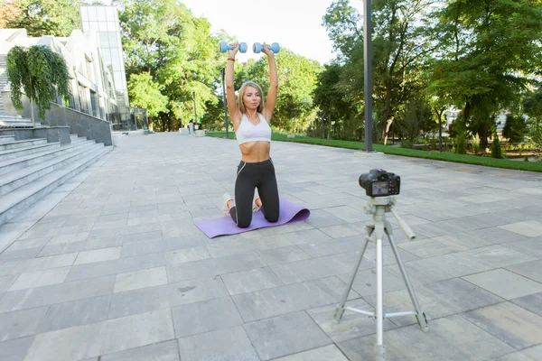 Fitness, sports and video blogging concept. Female sport blogger with camera on tripod recording outdoor fitness training with dumbbells