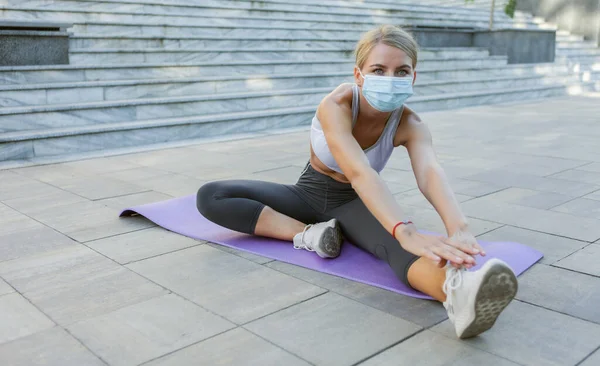 Slim fit woman in medical mask doing leg stretching on mat outdoors. Fitness workout during covid-19.