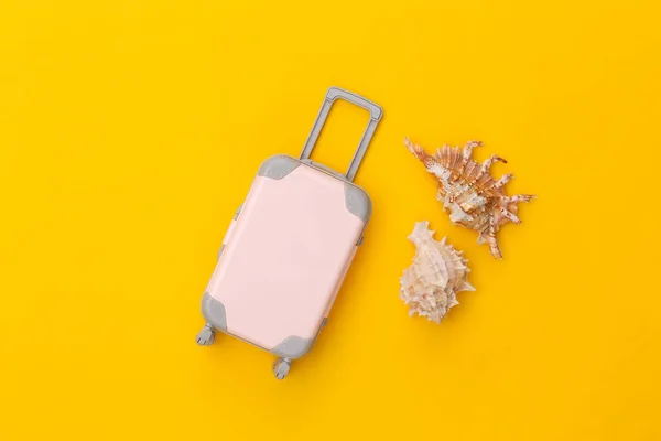 Travel concept. Mini plastic travel suitcase with seashell on yellow background. Minimal style. Top view, flat lay