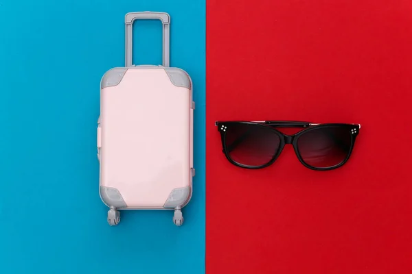 Travel concept. Mini plastic travel suitcase, sunglasses on red blue background. Minimal style. Top view. Flat lay