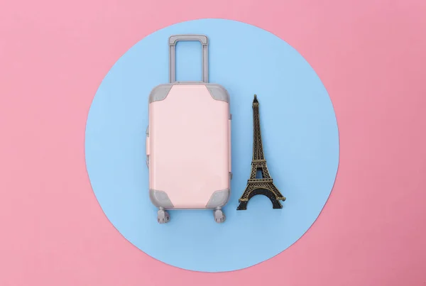 Travel minimalism. Mini plastic travel suitcase and statuette of the Eiffel Tower on pink background with blue circle. Minimal style. Top view, flat lay