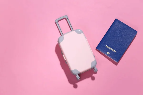 Travel minimalism. Mini plastic travel suitcase, passport on pink background with deep shadow. Minimal style. Top view, flat lay