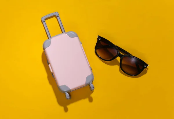 Travel minimalism. Mini plastic travel suitcase and sunglasses on yellow background with deep shadow. Minimal style. Top view, flat lay