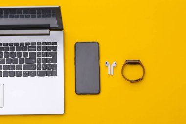 Modern aletler. Laptop, akıllı telefon, kablosuz kulaklık, sarı arka planda akıllı bilezik. Üst görünüm