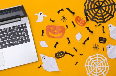 Sarı arka planda dizüstü bilgisayar ve kağıt kesikli Cadılar Bayramı dekoru. Üst Manzara. Düz yatıyordu