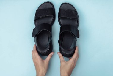 Dişi eller mavi arka planda siyah, modaya uygun sandaletleri tutuyor. Üst görünüm
