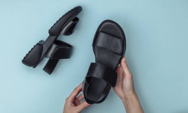 Dişi eller mavi arka planda siyah, modaya uygun sandaletleri tutuyor. Üst görünüm