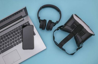 Modern aletlerin düz kompozisyonu. Laptop, video kask, kulaklık, mavi arka planda akıllı telefon. Üst görünüm