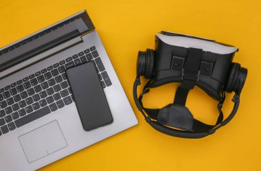 Modern aletlerin düz kompozisyonu. Laptop, video kask, kulaklık, sarı arka planda akıllı telefon. Üst görünüm