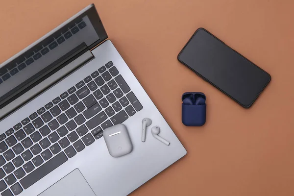Laptop ve kablosuz kulaklıklar, şarj kutusu ve kahverengi arka planda akıllı telefon. Üst görünüm